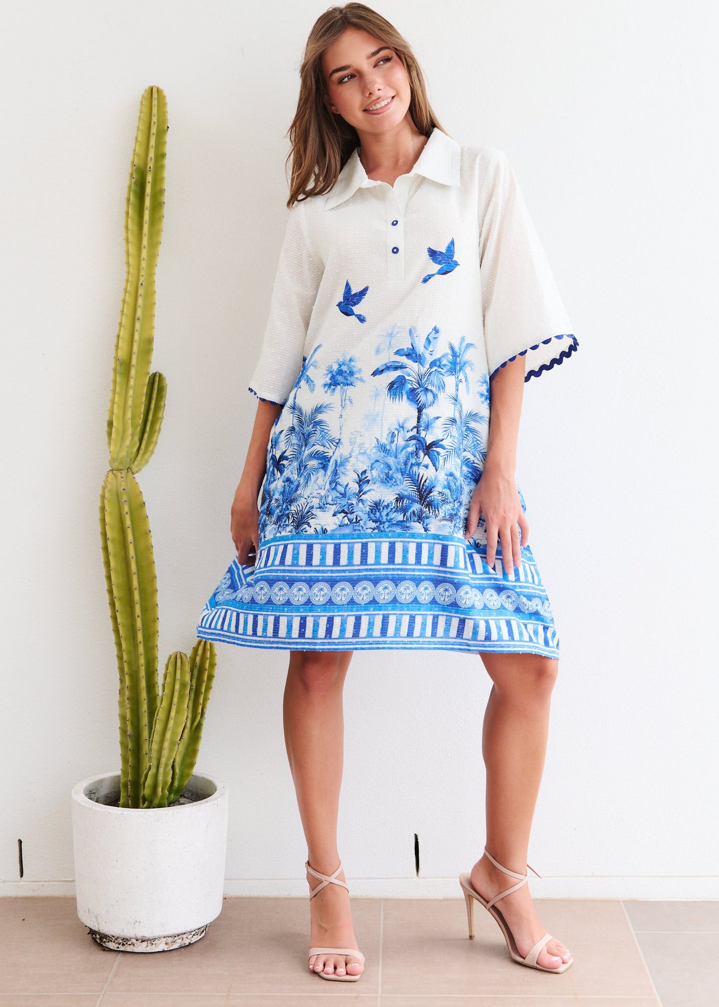 Woman in a floral dress standing next to a cactus plant indoors.