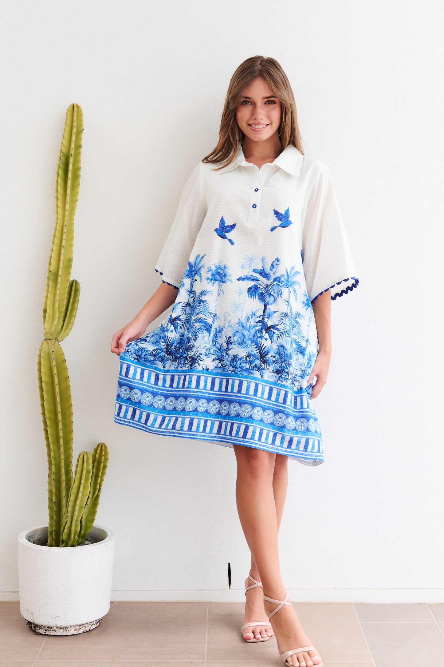 Woman wearing a white dress with blue floral patterns standing next to a potted cactus indoors.