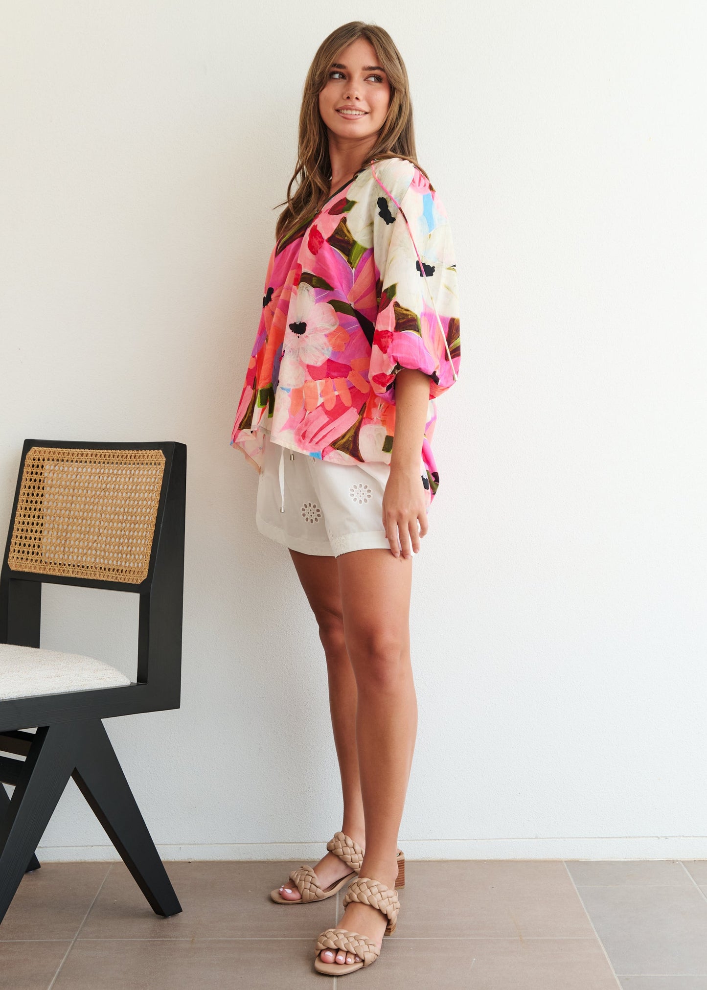Woman wearing a colorful floral blouse and white shorts standing next to a chair against a white wall.