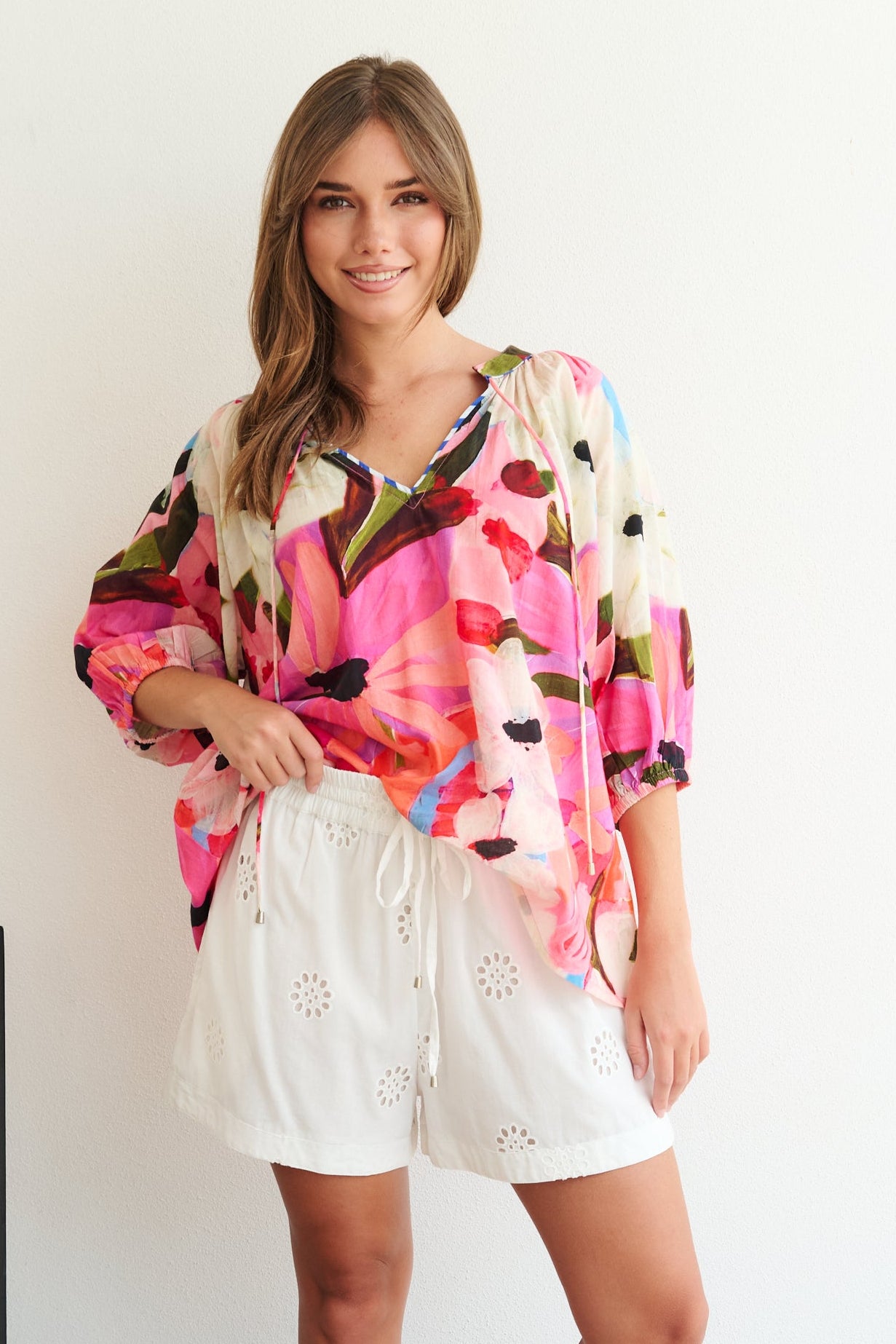 Woman wearing a colorful blouse and white shorts standing in a room with a chair and tiled floor.