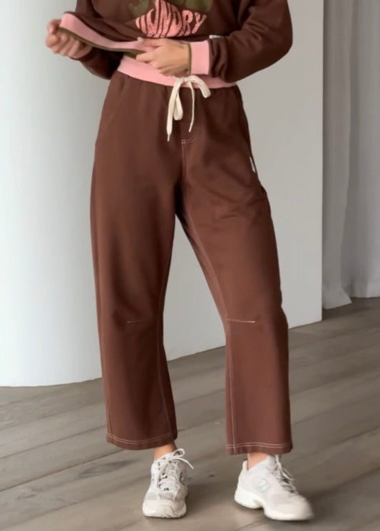 A woman modeling a brown two-piece lounge set consisting of a long-sleeve V-neck sweatshirt with a pink and green graphic print and matching brown sweatpants with a pink drawstring, standing in a modern room with light wood floors and white walls.