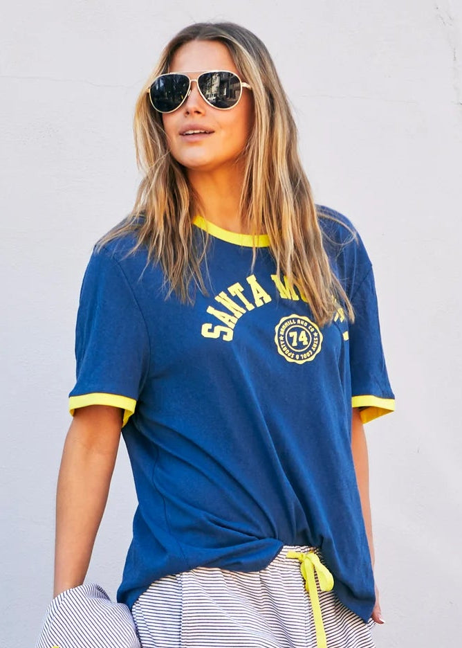 Woman wearing a blue sports jersey with yellow accents, gray shorts, and blue sneakers, standing against a white wall.