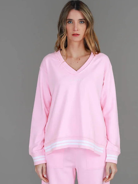 A woman modeling a light pink lounge set consisting of a long-sleeve V-neck sweatshirt with white stripe details and matching trackpants, paired with white sneakers, against a solid gray studio background.