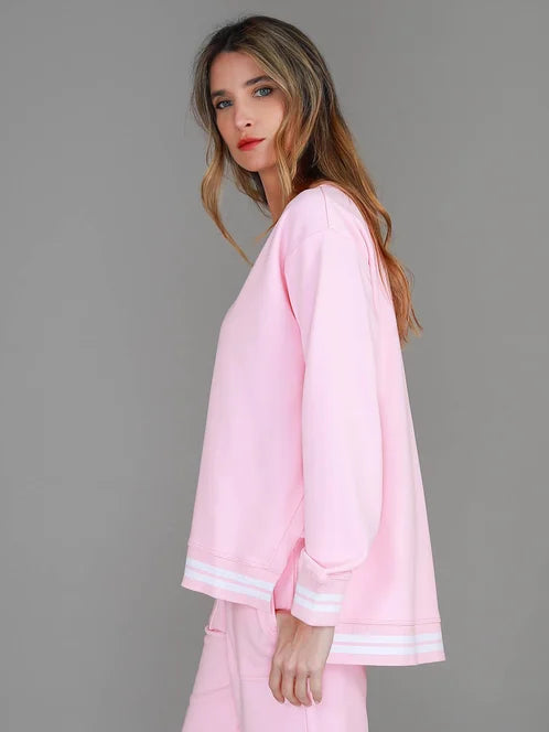 A woman modeling a light pink lounge set consisting of a long-sleeve sweatshirt and matching track pants, styled with white low-top sneakers against a solid gray studio background.
