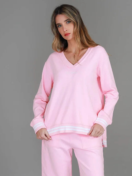 A woman modeling a light pink lounge set consisting of a long-sleeve V-neck sweatshirt with white striped cuffs and hem, paired with matching pink pants, against a solid gray studio background.