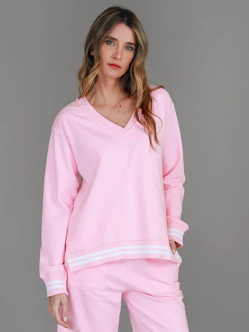 A woman modeling a light pink lounge set consisting of a long-sleeve V-neck sweatshirt with white stripes at the hem and matching track pants, paired with white sneakers, against a solid gray studio background.