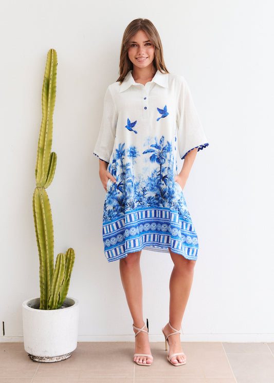 Woman wearing a white dress with blue floral patterns standing next to a cactus plant indoors.