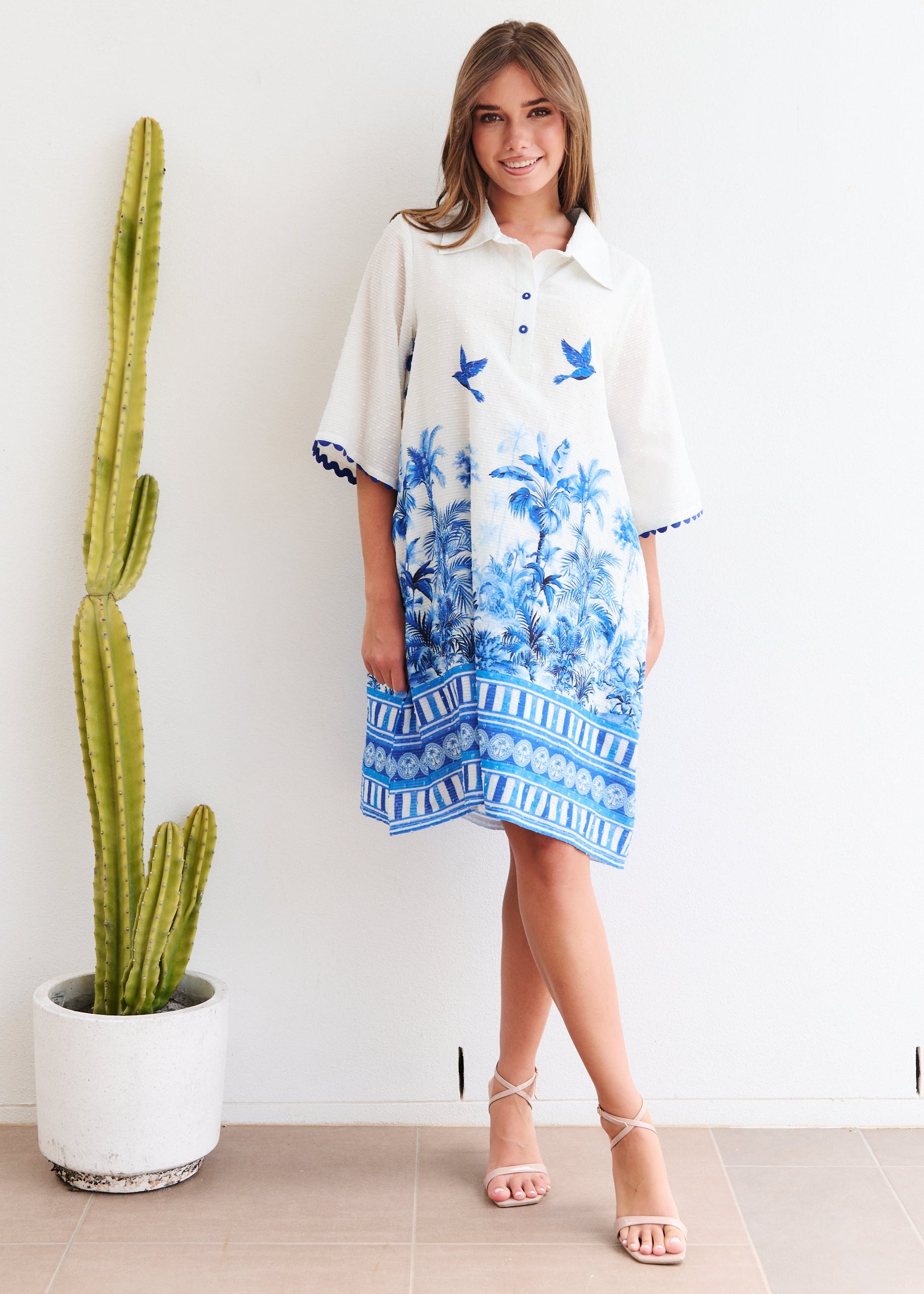 Woman wearing a floral dress standing next to a cactus plant indoors.