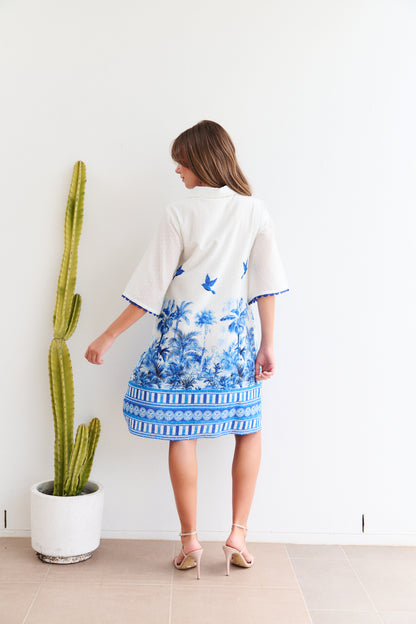 Woman wearing a white dress with blue floral patterns standing next to a cactus plant indoors.