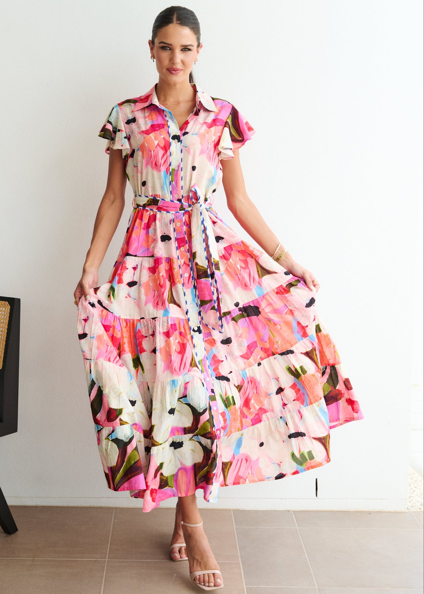 Woman wearing a colorful floral dress standing in a room with a chair and white wall.