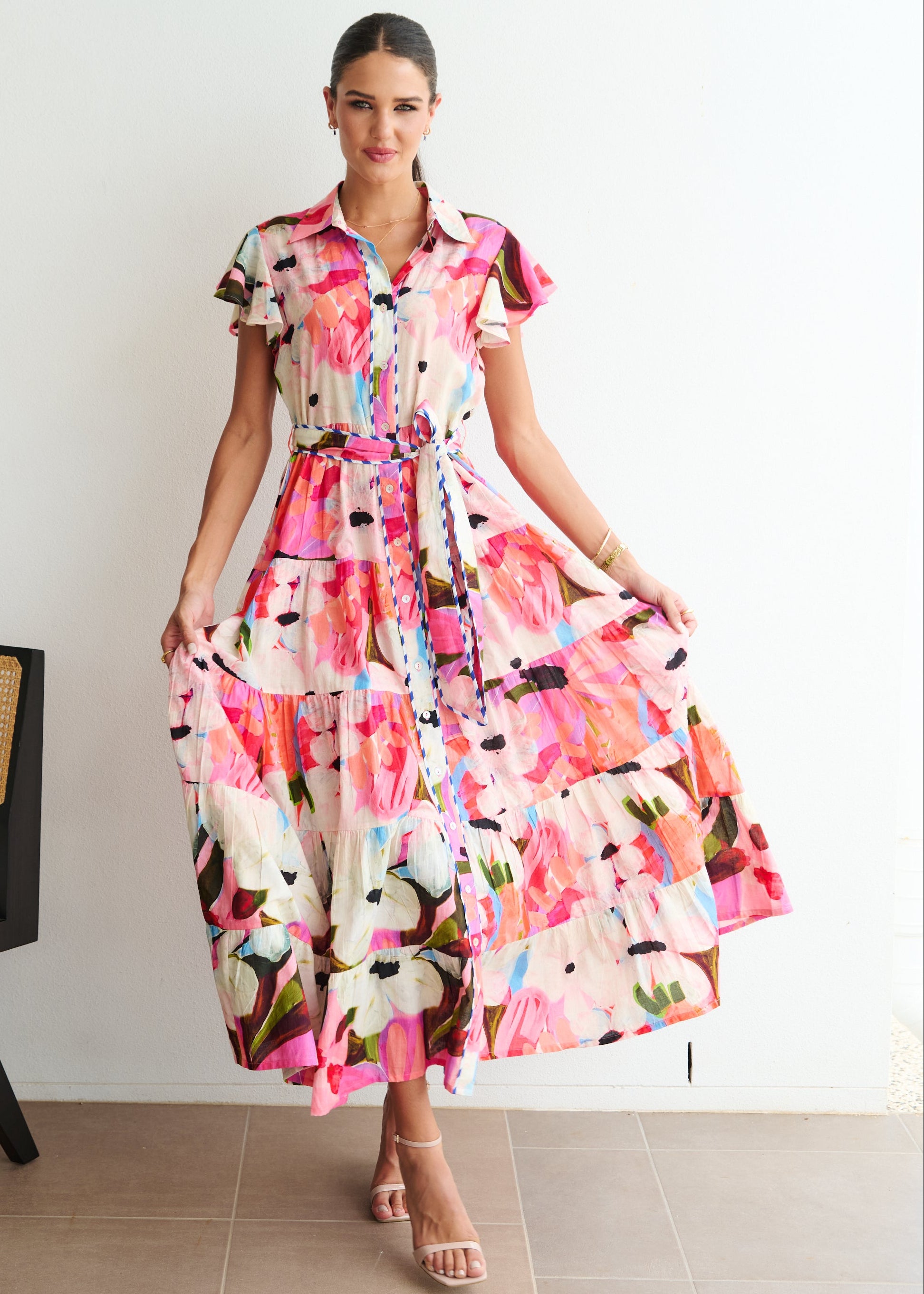 Woman wearing a colorful floral dress standing in a room with a chair and white wall.