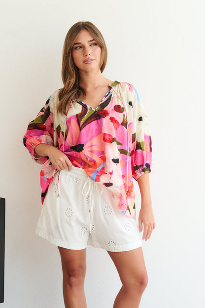 Woman wearing a colorful floral blouse and white shorts standing against a white wall.
