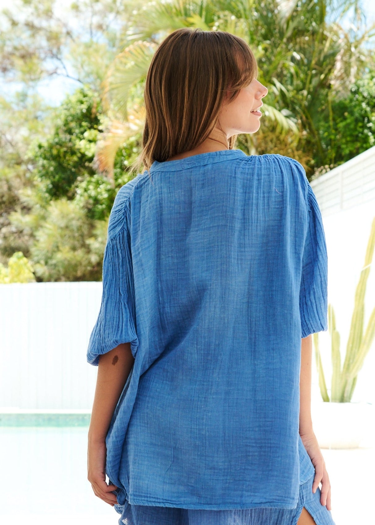 Woman wearing a blue cover-up standing in a bright room with greenery outside.
