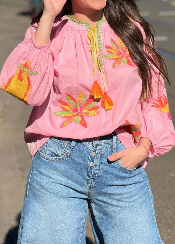 Woman wearing a pink blouse with floral embroidery and blue jeans on a street.