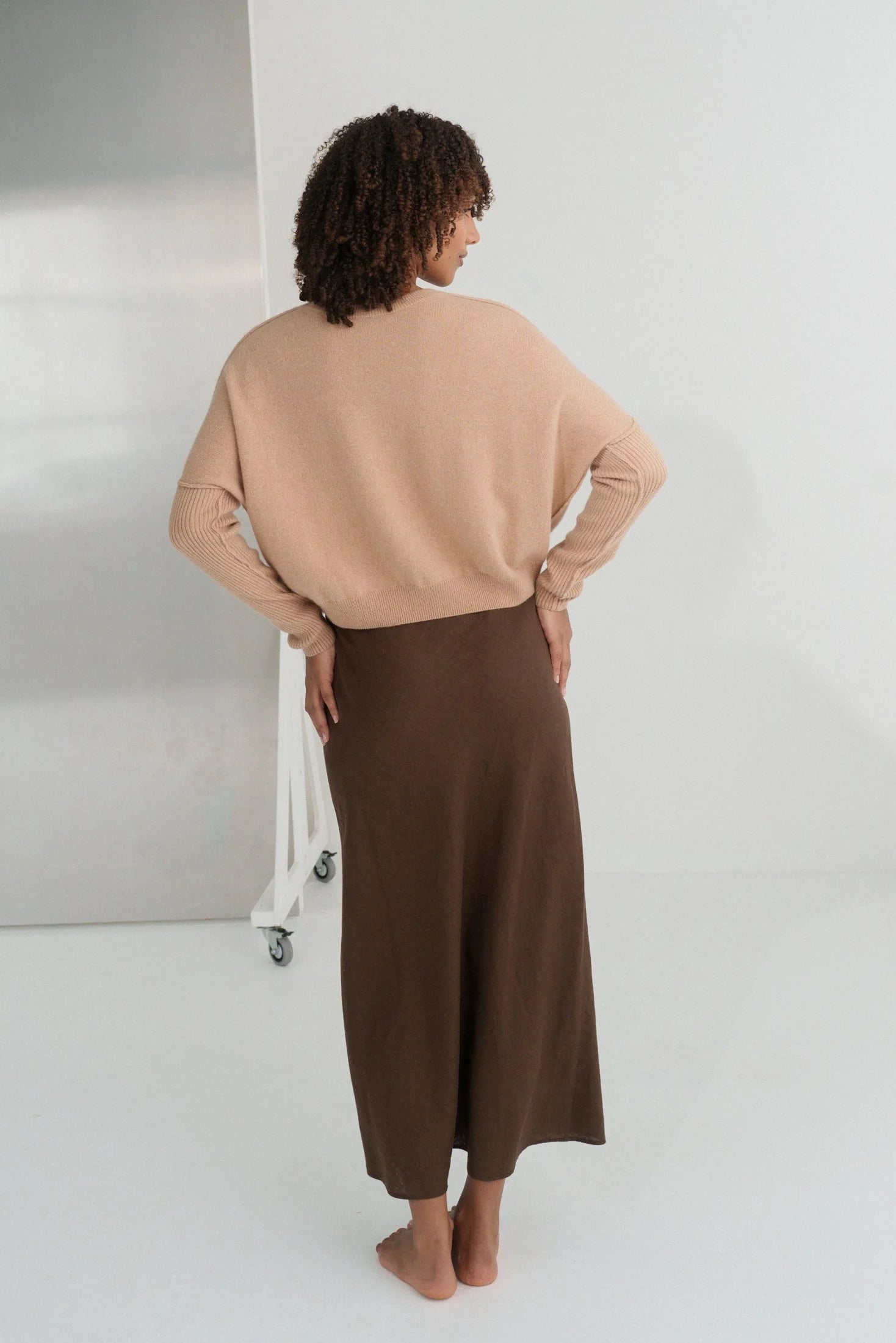 Woman wearing a beige cashmere sweater from the back and brown pants against a neutral background